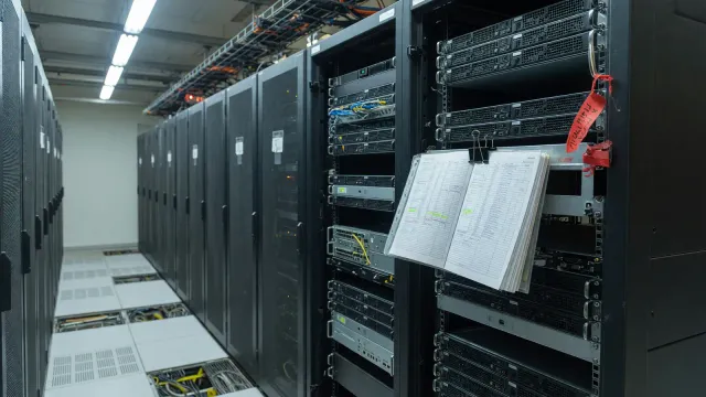 A server aisle in cool light, a rack pulled forward, a blank runbook on the rail and a red string tag on a handle.