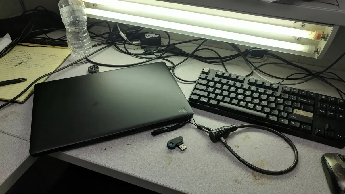 Linux workstation with a terminal and a closed laptop, cable lock on the desk, overhead office light, no neon.