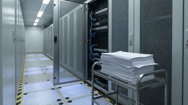 An open network-rack door and a face-down printout on a metal cart, cool white light, no vendor logos and no readable text.